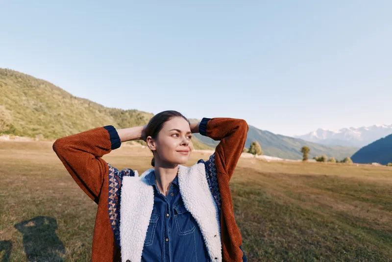Woman portrait outdoors in mountains meadow, relaxation and travel vibe. Casual young female in jacket and denim smiles with hands behind head enjoying scenic countryside and sky.
