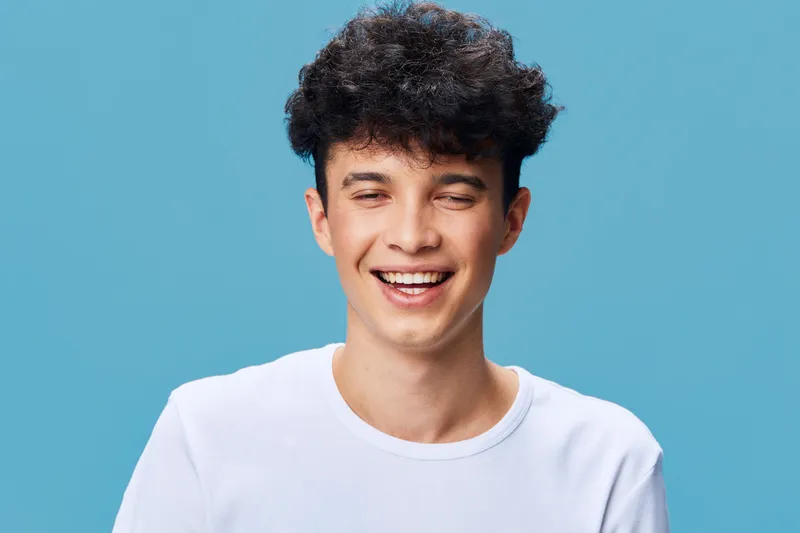 smiling man with curly hair wearing white shirt against blue background expressing happiness and joy in casual portrait bright lighting