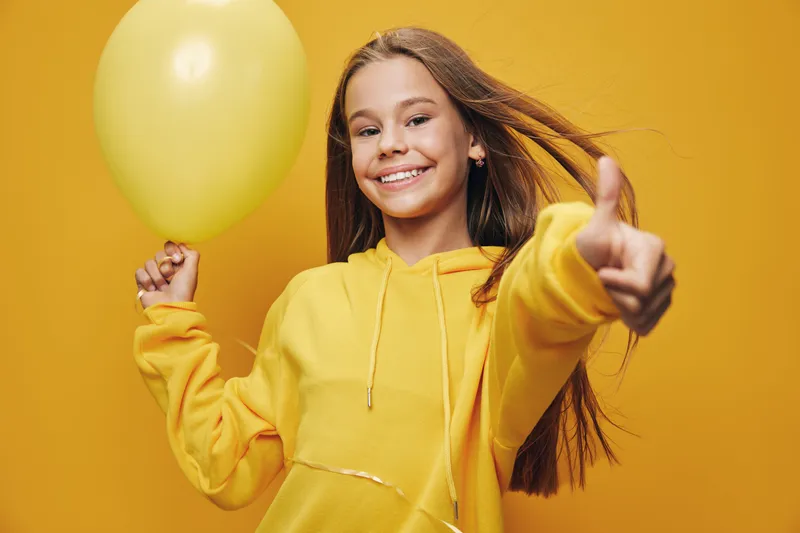 Girl smiling and giving thumbs up while holding yellow balloon against yellow background. Child with long hair in yellow hoodie enjoys celebration mood with happiness.