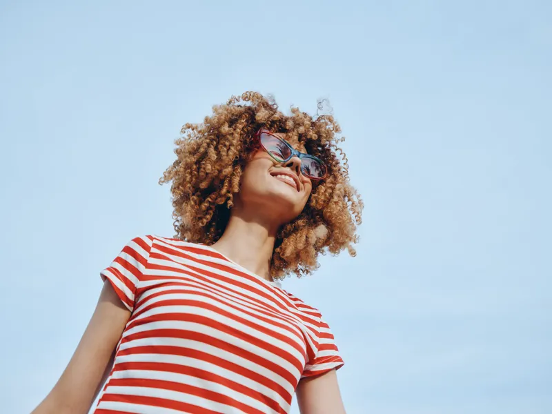 A stylish portrait of a smiling person with curly hair wearing a red and white striped tee and sunglasses, standing under a clear blue sky on a sunny day.