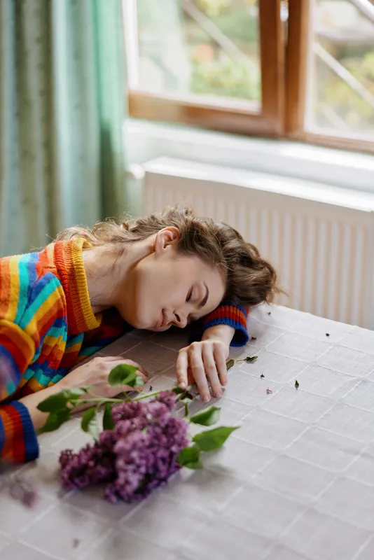 Relaxed woman at home lies on a table beside lilac flowers, wearing a colorful striped sweater, daylight creates a calm intimate mood, candid lifestyle moment indoors