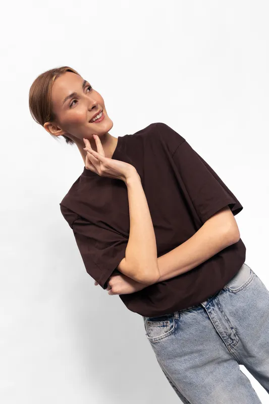 Young woman in casual brown t-shirt and blue jeans looks thoughtfully to side, posing with hand near face on white background. Concept for fashion, lifestyle, and natural beauty.