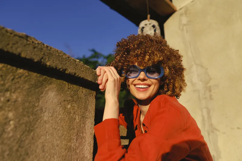Young woman with curly hair in a red sweater smiles warmly while leaning on a sunlit concrete wall, creating a candid moment of casual confidence and everyday wellness on a bright day