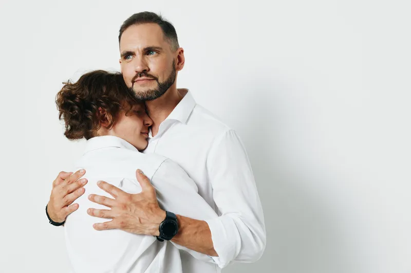 An executive man in a smart shirt embraces a colleague, conveying support and empathy. The scene uses a solid colored backdrop to isolate the professional emotion and connection.