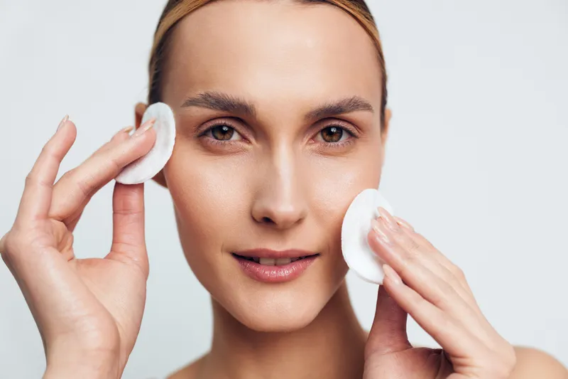 Close-up of woman with clear skin cleansing face with cotton pads, skincare routine on white background. Concept of beauty, hygiene, wellness, and self-care with soft natural lighting.