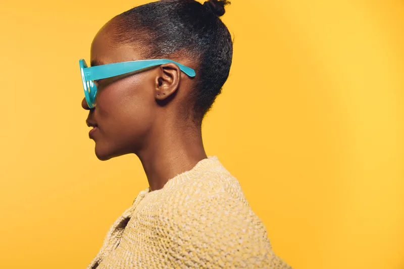 Profile of African woman with dark skin and blue sunglasses on yellow background. She wears a beige knit sweater, showcasing modern fashion and confident style concept.