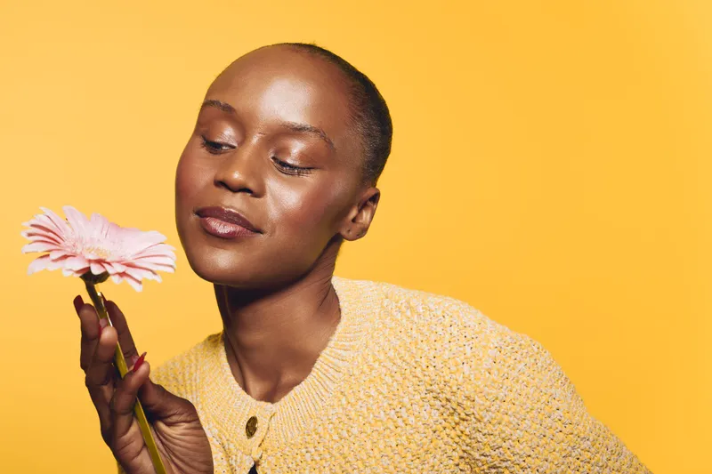 Portrait of a dark skinned African woman holding a pink flower and wearing a yellow knitted sweater. Soft studio lighting highlights her serene expression on a yellow background.