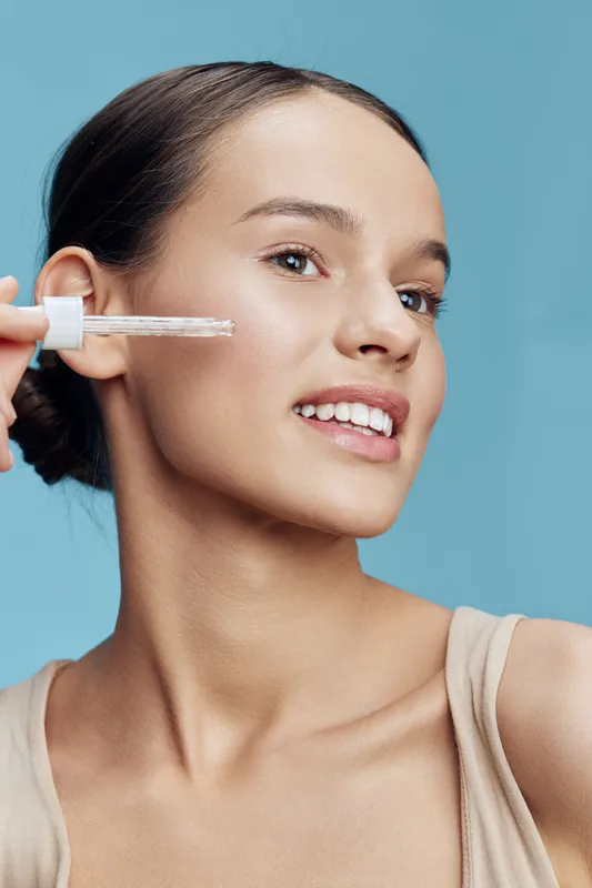 Young woman applying serum to her skin, showcasing a radiant smile against a soft blue background, emphasizing beauty and skincare