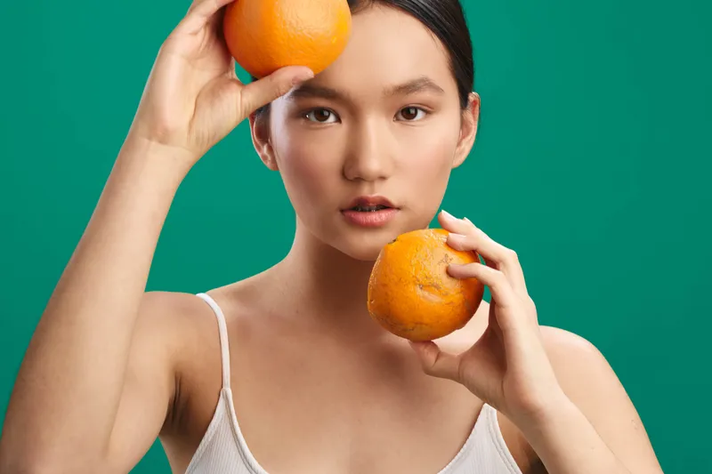 Asian woman holds oranges against a vibrant green backdrop, posing with a calm expression. The image emphasizes freshness, simplicity, and a modern lifestyle aesthetic suitable for wellness and