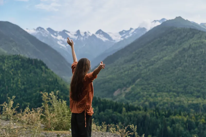 Woman stands with arms raised in a vast mountain landscape, celebrating nature and freedom as snowy peaks rise and a lush valley unfolds.