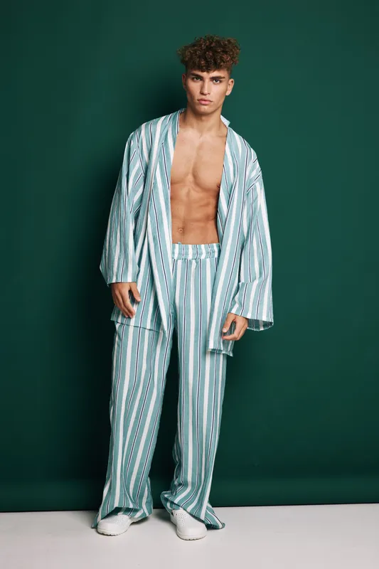 Man with bare chest wearing striped loose jacket and pants standing on beige floor against dark green background with casual modern clothing and stylish look expressing confidence.