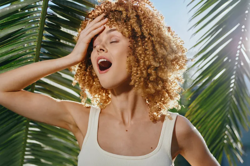 smiling woman with curly hair stands in sunlight near tropical palm leaves, wearing white tank top and expressing joy in bright outdoor summer setting.