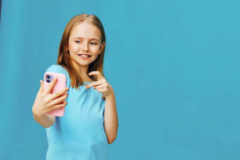 Happy young girl taking a selfie with a smartphone against a blue background, wearing a light blue shirt, showcasing joy and playfulness in a modern, vibrant style