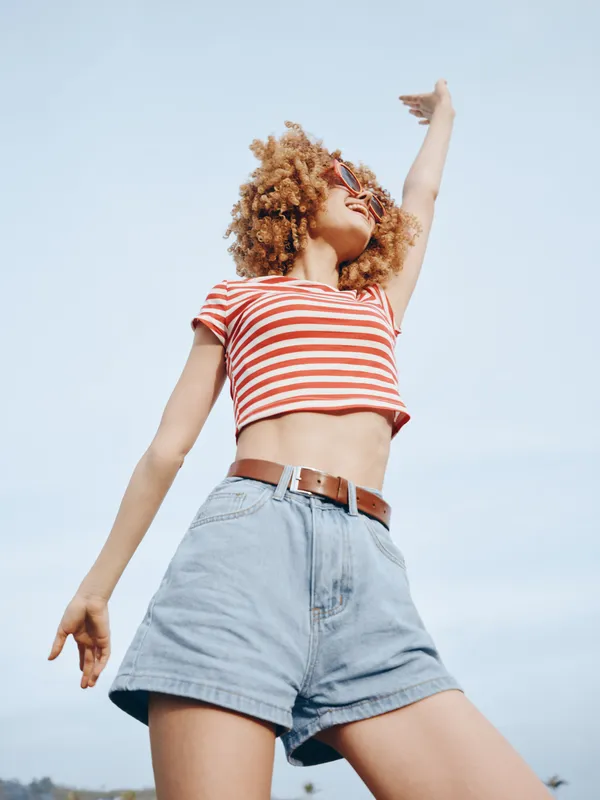 Young woman enjoys a sunny day outdoors, wearing a striped crop top and high waist denim shorts. She stretches arm upward, smiling confidently against a clear blue sky.