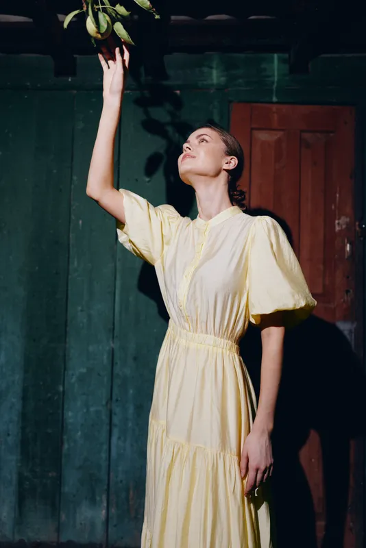 Woman in a yellow dress reaches toward a plant in a dim indoor space, standing beside a wooden doorway, creating a vintage portrait with soft lighting and calm mood