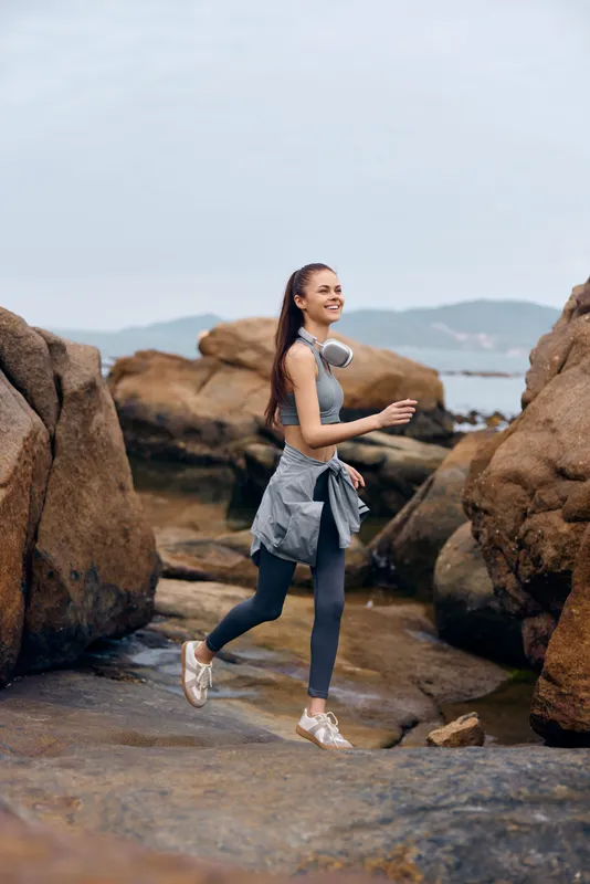 Young athletic woman jogging on rocky beach, wearing sportswear, smiling and enjoying nature, capturing the essence of a healthy lifestyle and fitness