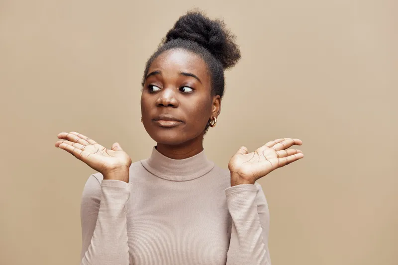 Confused African American woman with neutral expression showing hands in a shrug gesture, wearing a beige turtleneck, on a soft brown background