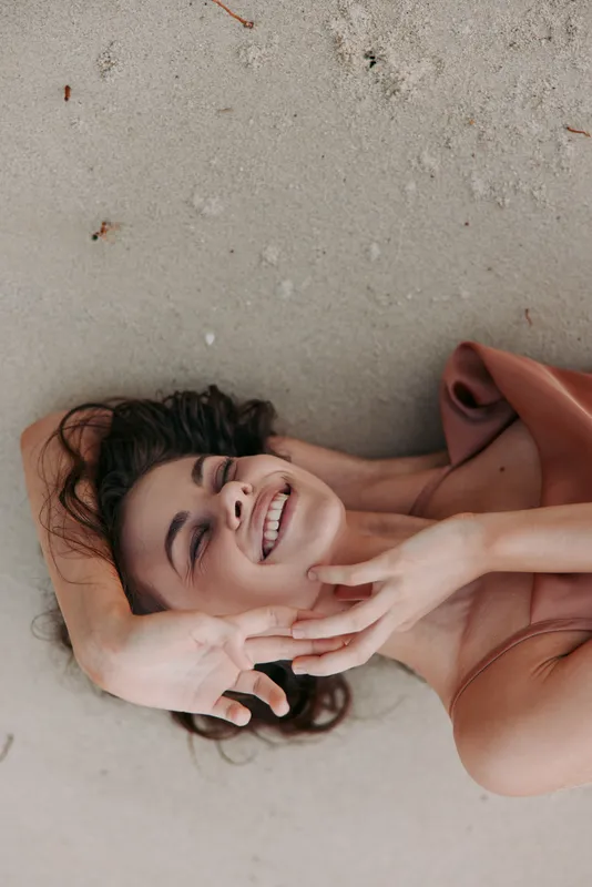 A joyful woman lies on soft sand, smiling with closed eyes, head tilted, arms resting, wearing a flowing dress. Warm sunlight highlights a relaxed mood and carefree moment on the shore.
