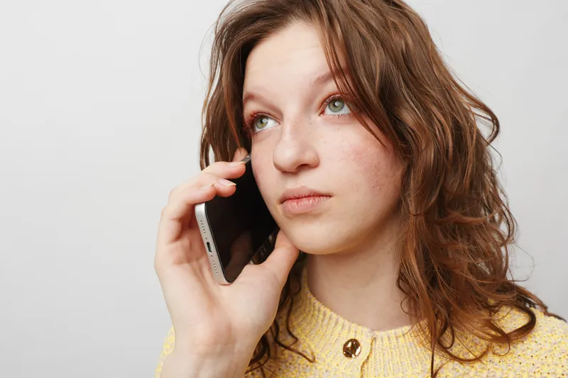 Young woman with curly brown hair wearing yellow sweater talks on smartphone looking away thoughtful. Concept of communication, technology, and casual lifestyle in studio setting.