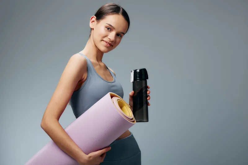 Fitness, sport and woman in athletic wear holding a yoga mat and water bottle for workout, yoga and training in a clean studio setting, showcasing wellness and an active lifestyle.