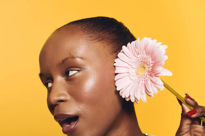 Close-up of a dark skinned African woman with short hair holding a pink flower near her ear. Bright yellow background creates a vibrant and fresh mood focused on beauty and nature.
