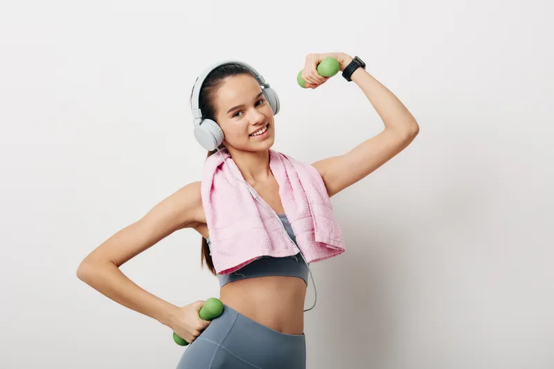 Woman fitness portrait of a young woman lifting green dumbbells, wearing headphones and a pink towel over her shoulders in sports bra and leggings, smiling against a clean white background.