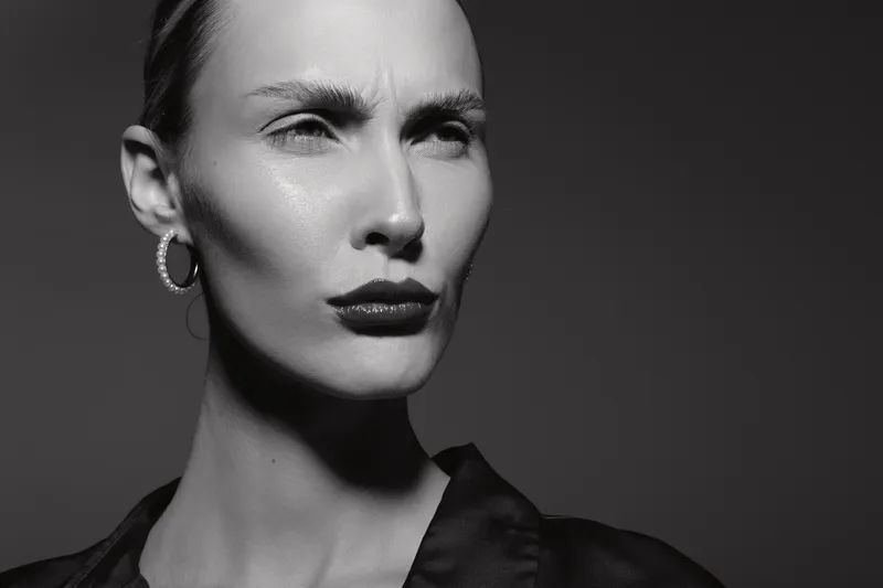 Close up portrait of a serious caucasian woman with pale skin and slicked hair wearing hoop earrings and dark lipstick in monochrome.