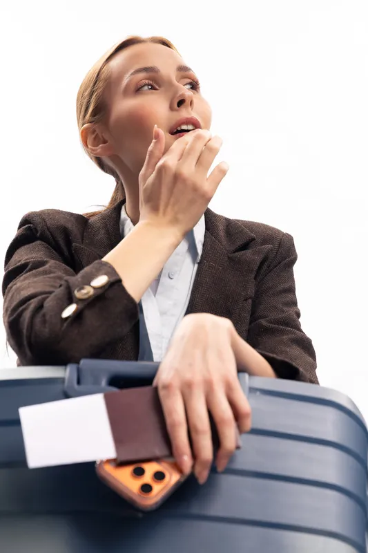 Young woman in brown blazer holds blue suitcase and smartphone with boarding pass, looks away with thoughtful expression. Concept of travel, business trip, and planning ahead.
