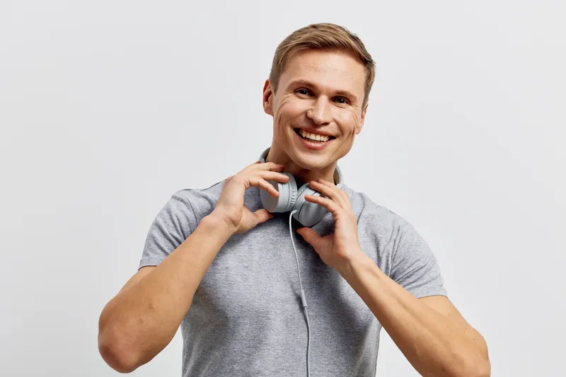 Smiling happy young man wearing casual gray t-shirt holding headphones near neck pose isolated on plain white background. Studio portrait of cheerful male model with friendly expression. People lifest