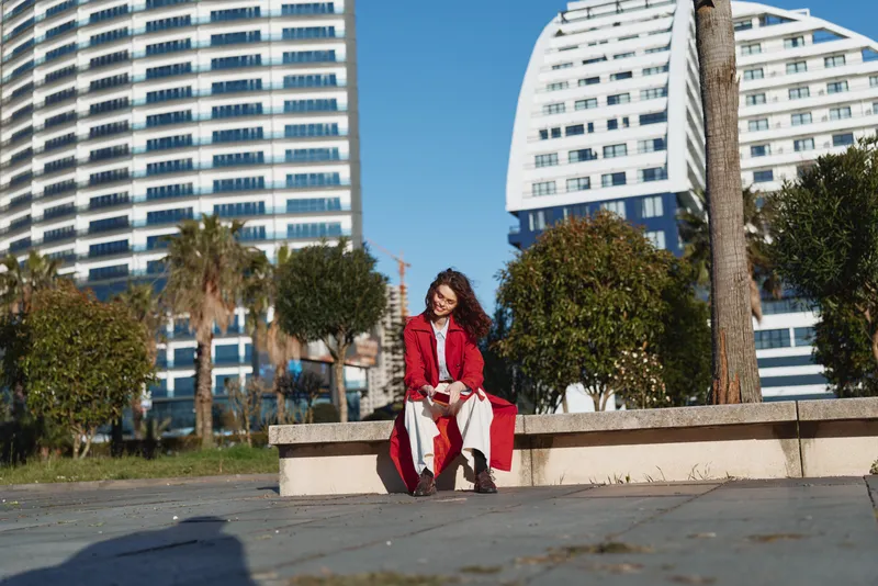 Young woman in red coat sitting on bench outdoors in urban park, sunny day, modern buildings background, relaxed and thoughtful mood