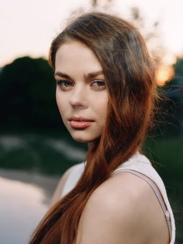 Beautiful young woman with long hair, soft expression, wearing casual light clothing, sunset background reflecting serenity and natural beauty.