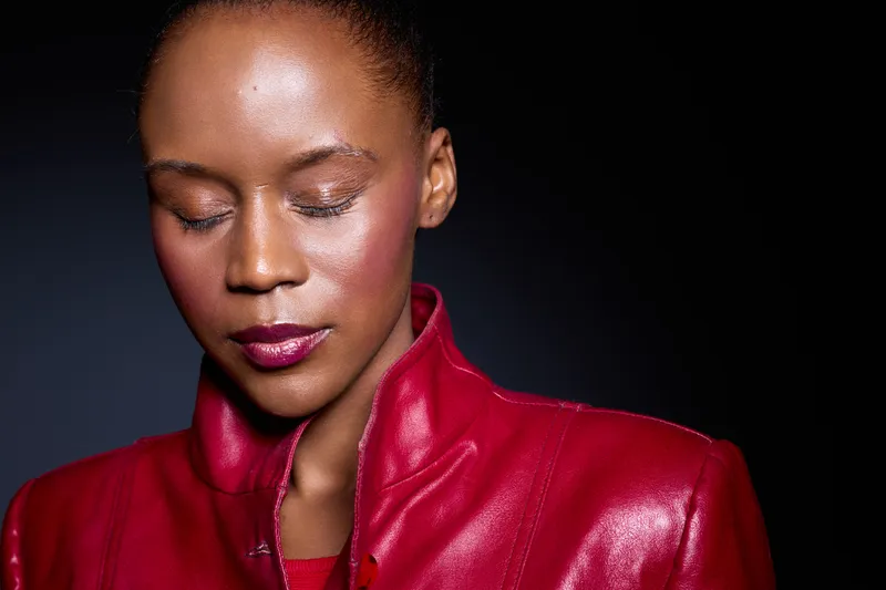 Portrait of a thoughtful black woman with dark skin wearing a shiny red leather jacket, eyes closed against a dark background. Concept of fashion, elegance, and introspection.