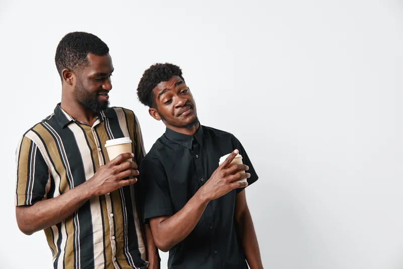 Two African American men with dark skin holding coffee cups and smiling, casual clothing, friendly interaction and relaxed mood on white background.