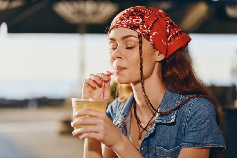 Lifestyle woman enjoying a refreshing drink in modern boho-western denim street style with film color tones, relaxed mood, sunny outdoor setting, and casual summer vibe.