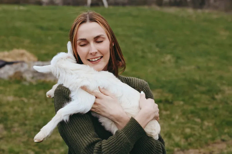 Young woman with red hair happily hugs a white baby goat outdoors in a green field, enjoying a tender moment with the animal in casual autumn clothing.