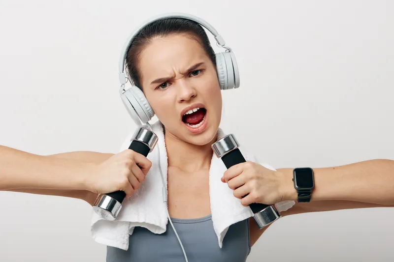 Fitness woman in athletic wear lifts dumbbells, wears headphones and a towel, showing intense effort during a focused workout in a clean studio setting.