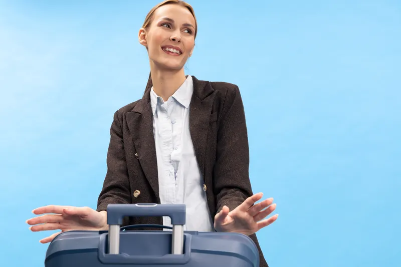 Young businesswoman with suitcase stands against blue background smiling, ready for travel or business trip. Concept of travel, work, confidence, and preparation for journey.