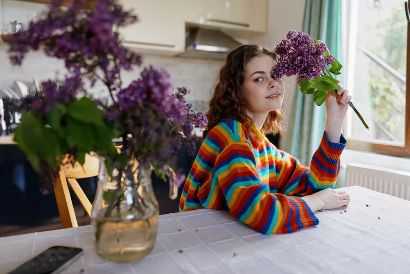 A young woman in a striped sweater sits at a dining table indoors, smelling a lilac bouquet held close by a vase of flowers, in a bright cozy home setting with natural light on her face