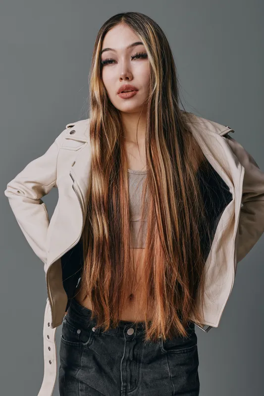 Young woman with long straight hair and beige jacket posing confidently against grey background. Casual fashion style with modern hairstyle and neutral expression.