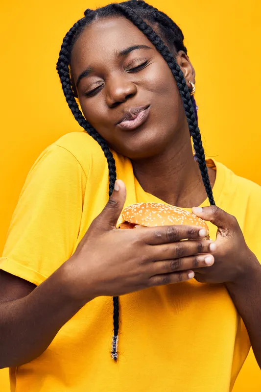 A joyful young woman of African descent wearing a bright yellow t shirt, holding a fresh burger against a vibrant yellow background, radiating happiness and excitement