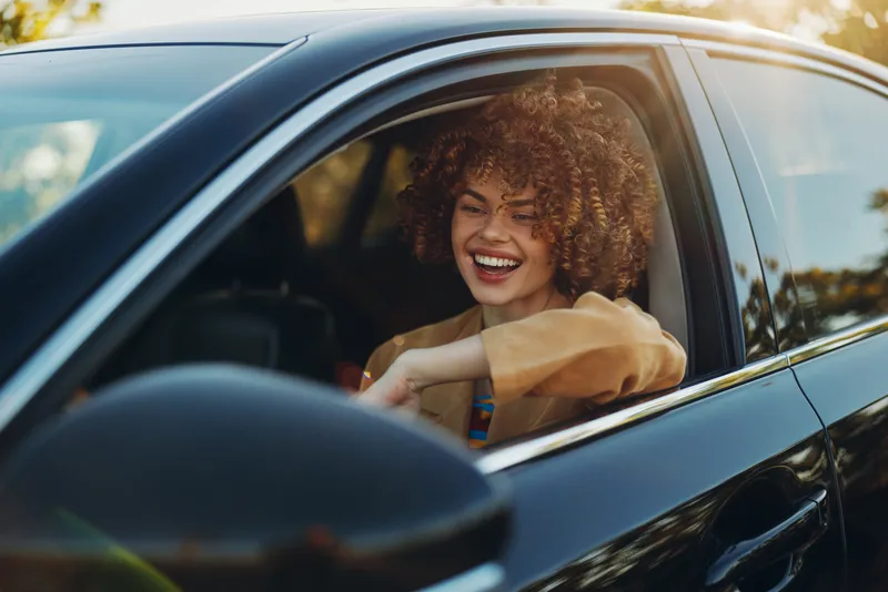 A joyful woman sits in a car window, smiling broadly as sunlight highlights her curly hair. Casual everyday moment captured with retro film color warmth.