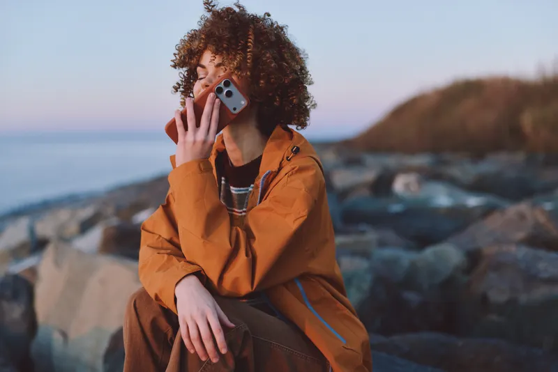 Woman lifestyle at seaside, seated on rocks, holding a smartphone near her ear, curly hair, orange jacket, outdoors by the ocean and waves, casual travel mood, soft daylight, relaxed calm face