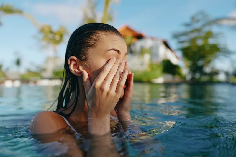 Woman in a clear pool rinses her face with water, hands on cheeks, a refreshing summer moment, tropical resort backdrop, relaxed vibe, swimwear, sunlit scene
