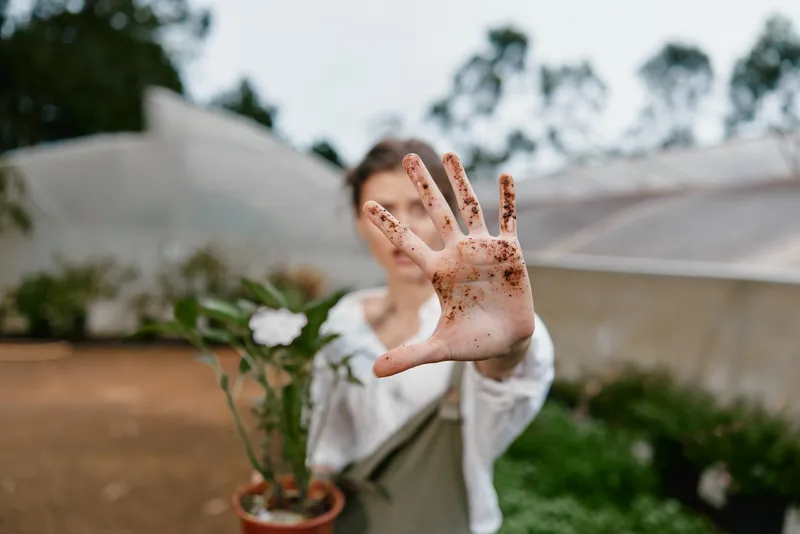 A young woman with soil stained hands showcases her love for gardening in a vibrant, green greenhouse, emphasizing growth, nature, and sustainability