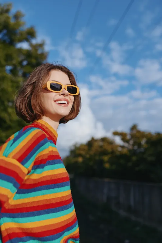 Woman smile sunglasses colorful sweater outdoors portrait happy short hair on sunny day, candid side view with blue sky, trees and bright retro knitwear showing confidence and joy