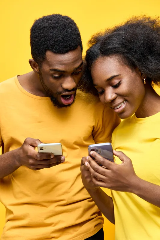 Young African couple joyfully interacting with smartphones against a bright yellow background, showcasing excitement and modern lifestyle choices