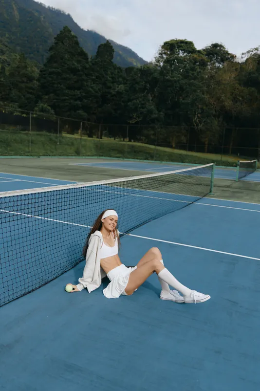Young female athlete relaxing on a tennis court, showcasing sporty attire and a joyful expression against a lush, green background