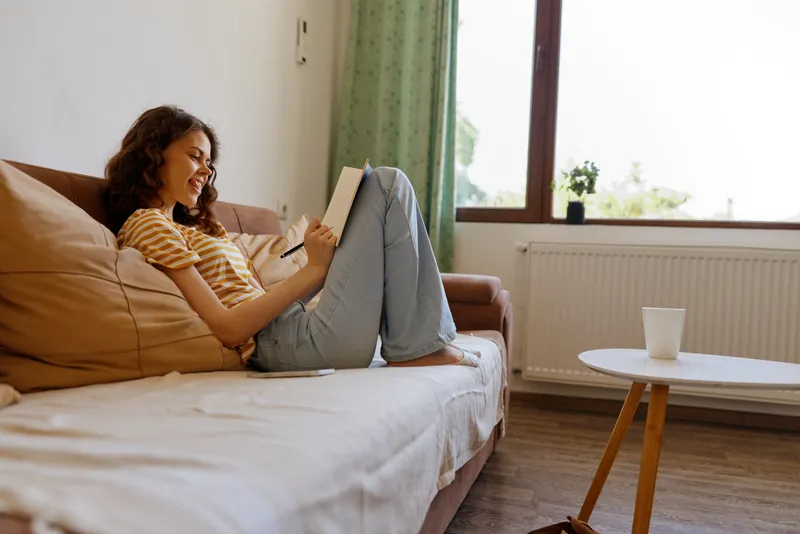 Woman relaxing at home on a sofa, reading on a tablet, casual clothing, cozy interior with sunlight, leisure mood, everyday moment