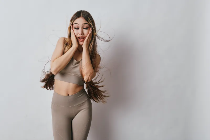 Woman in fitness clothing with long hair expressing surprise or shock on plain light background. Studio portrait of female model with hands on face and braid hairstyle.