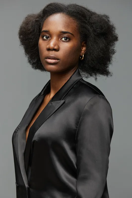 Portrait of a confident Black woman wearing a sleek black satin blazer with natural hair, showcasing a serious expression against a subtle gray background